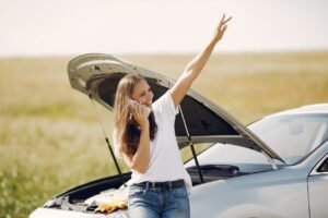 Cheerful young female driver in white t shirt and jeans speaking on phone and looking away with smile while leaning on broken car with open hood and showing greeting gesture with raised hand
